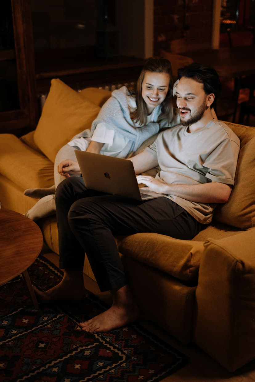 Young couple sitting together on a golden-yellow couch in a warmly lit living room, smiling while looking at a MacBook laptop. The woman is leaning against the man as they engage with financial management tools, representing stress-free money management in a comfortable home setting.