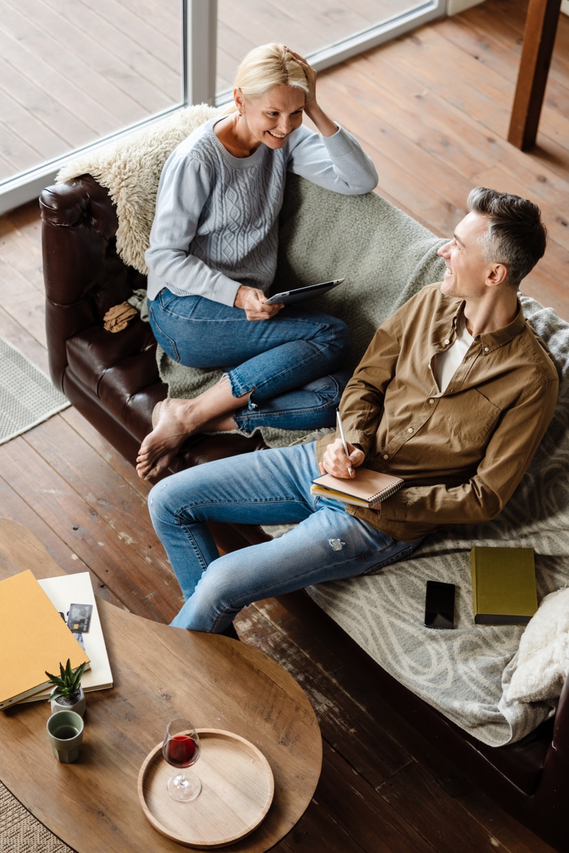Young couple sitting together on a golden-yellow couch in a warmly lit living room, smiling while looking at a MacBook laptop. The woman is leaning against the man as they engage with financial management tools, representing stress-free money management in a comfortable home setting.